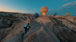 man riding a bicycle on a mountain trail with hot air balloons in the background 