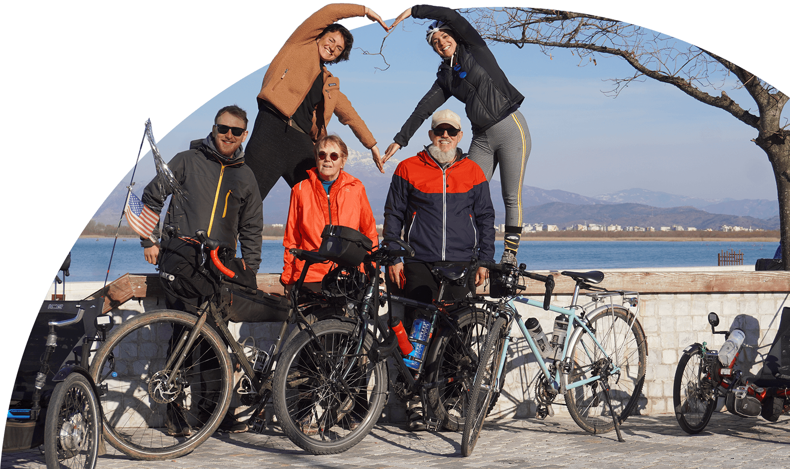 5 cyclists standing by a lake with 2 of them making a heart together with their arms